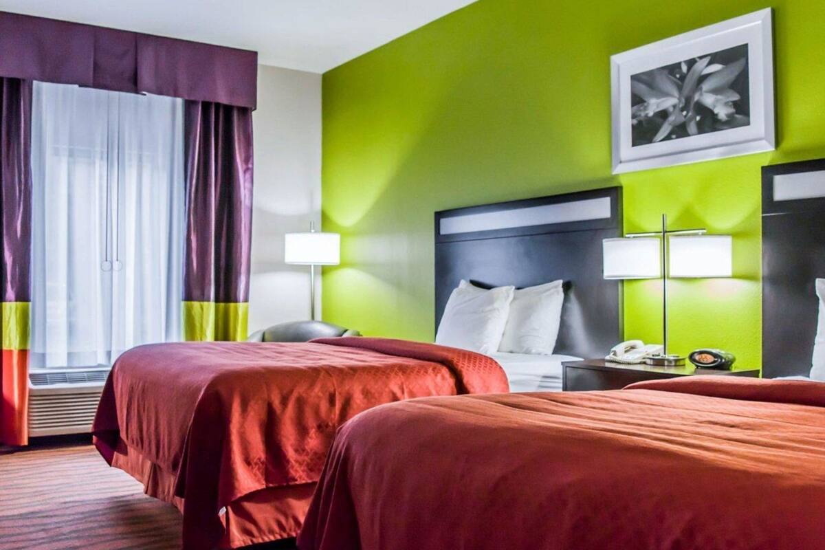 A hotel room with two beds, a large window with curtains, a green accent wall, and a framed picture on the wall.