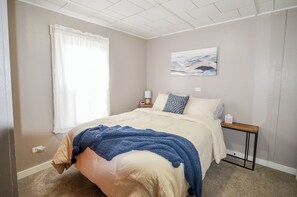 Una camera da letto con un letto, un comodino, una lampada, un quadro appeso e una finestra con tende.
