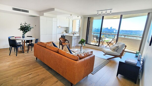 Un soggiorno moderno con un divano in pelle marrone, un tavolo da pranzo con sedie, una zona cucina e un balcone con vista sulla città.