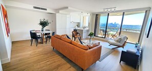 Un salon moderne avec un canapé en cuir marron, une table à manger avec des chaises, un coin cuisine et un balcon offrant une vue sur la ville.