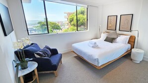 Una camera d'albergo moderna con una grande finestra, un letto con lenzuola bianche, un divano blu e un tavolino con un vaso di fiori.