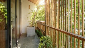Een balkon met een bamboe hek, een betonnen plantbak met een boom, en uitzicht op groen.