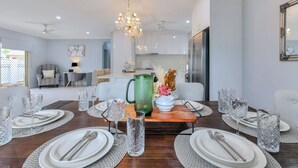A dining area with a wooden table set for six, surrounded by chairs. There is a chandelier above the table, and a kitchen is visible in the background. - The OC Home: Centrally Located, Home Away Home (Coconut Grove)