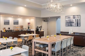 Ein Essbereich eines Hotels mit einem zentralen Holztisch, umgeben von Stühlen, und einem Küchenbereich im Hintergrund.