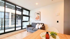 A modern living room with a wooden dining table, a grey sofa, and a large window with blinds.