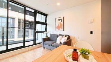 A modern living room with a wooden dining table, a grey sofa, and a large window with blinds.