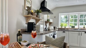A modern kitchen with a dining area, featuring a round table, a counter with a coffee machine, and a window with a view of greenery. - Drovers Run - Beaminster, Dorset (Chard)