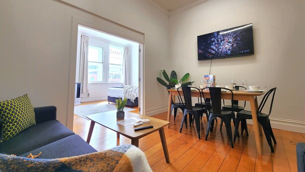 A modern living room with a sofa, coffee table, and dining area with chairs and a table. A TV is mounted on the wall, and a window provides natural light. - U Suites on Cuba (Wellington)