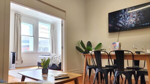 A dining area with a wooden table, black chairs, and a potted plant. A television mounted on the wall displays a cityscape. - U Suites on Cuba (Wellington)