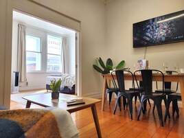 A dining area with a wooden table, black chairs, and a potted plant. A television mounted on the wall displays a cityscape.