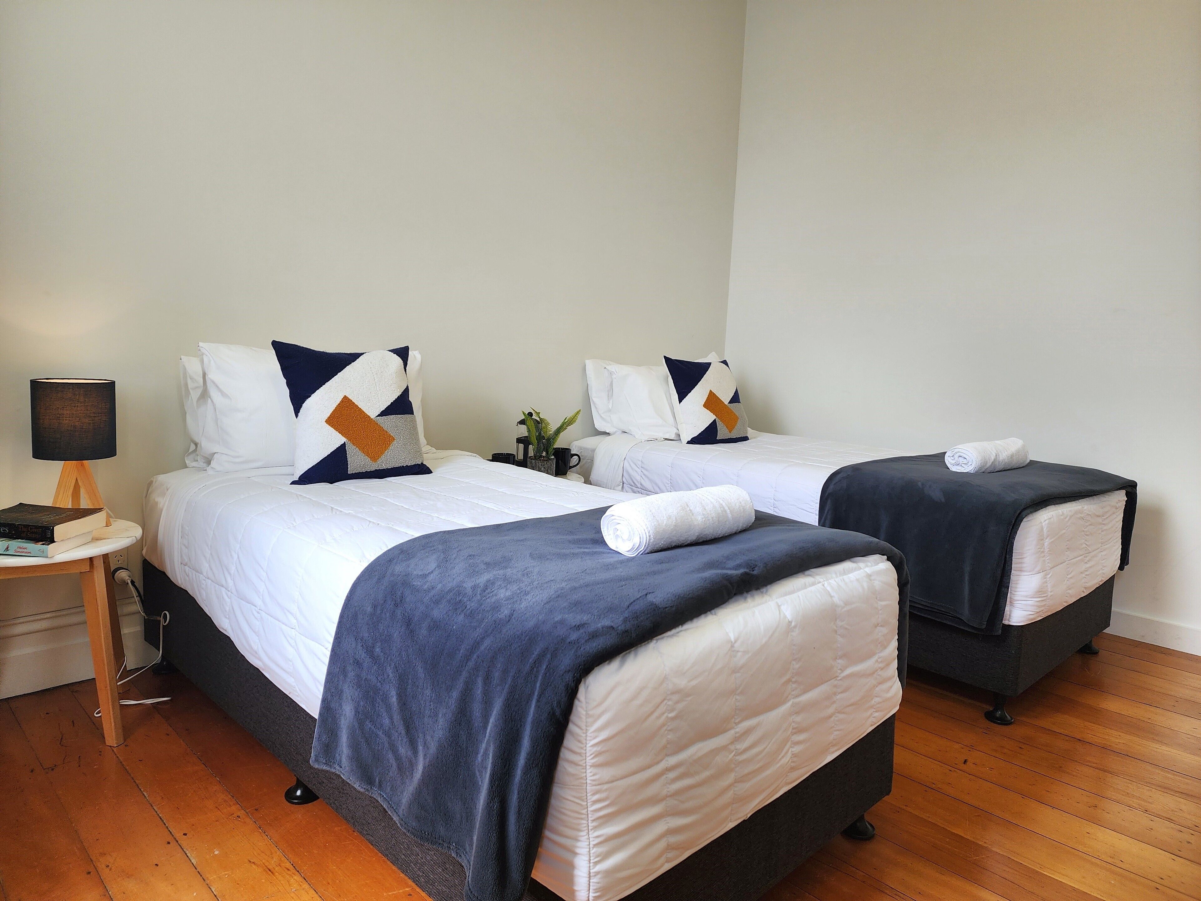 A hotel room with two beds, each with a white duvet and navy blue throw blankets. There is a bedside table with a lamp and a small plant.