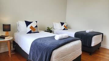 A hotel room with two beds, each with a white duvet and navy blue throw blankets. There is a bedside table with a lamp and a small plant.