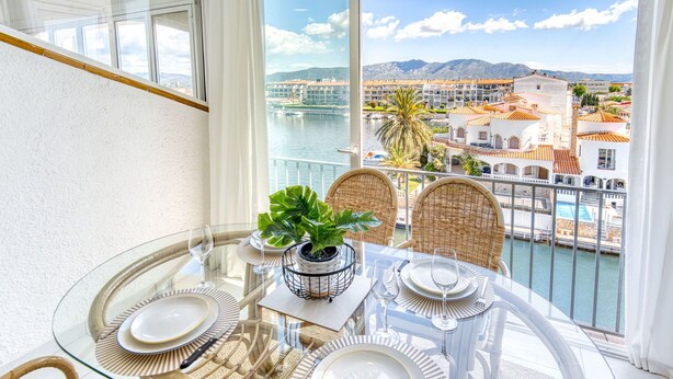 Un balcón con una mesa de vidrio dispuesta para comer, sillas de mimbre y vistas a un pueblo costero y montañas.