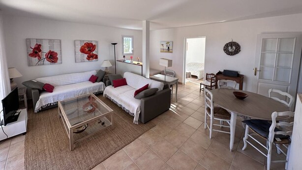 A spacious living area with a sofa set, a glass coffee table, and a dining table with chairs. There is a television, a lamp, and artwork on the walls.