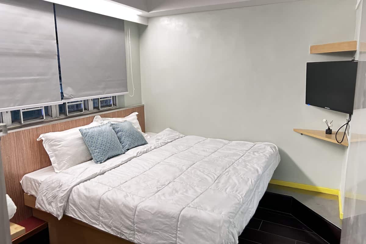 A neatly made bed with white bedding and two pillows, a bedside table with a red cushion, a mounted TV on a wooden shelf, and a window with blinds.