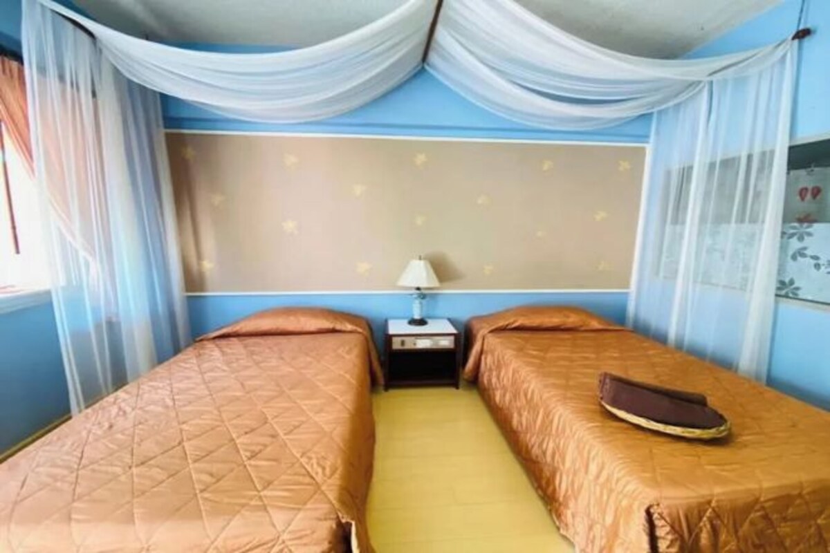 A room with two beds, a nightstand, and a window with sheer curtains.
