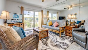 A spacious living room with a large window, a ceiling fan, and a flat-screen TV displaying 'tripvacations key west'.