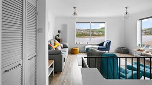 A spacious living room with a view of a lake, a grey sofa, a blue armchair, and a wooden coffee table. - Swan's Nest - River view, Close to beach, Garden (Torcross)