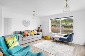A modern living room with a grey sofa, colorful cushions, a teal armchair, and a large window offering a view of a lake and houses.