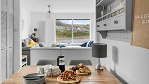 A modern kitchen with a wooden table set for breakfast, a coffee maker, and a view of houses through the window. - Swan's Nest - River view, Close to beach, Garden (Torcross)