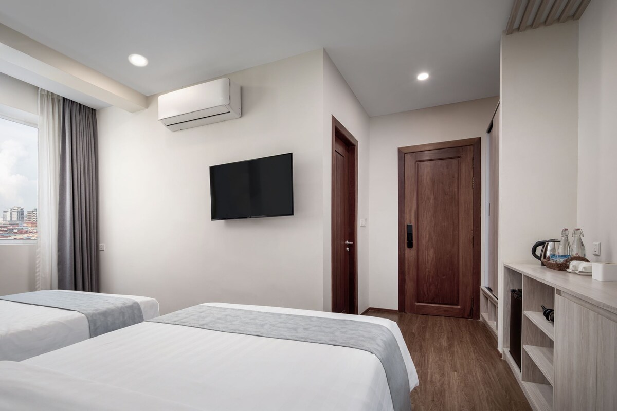 A hotel room with two beds, a TV, an air conditioning unit, a wooden door, and a small kitchenette with a coffee maker and bottles.