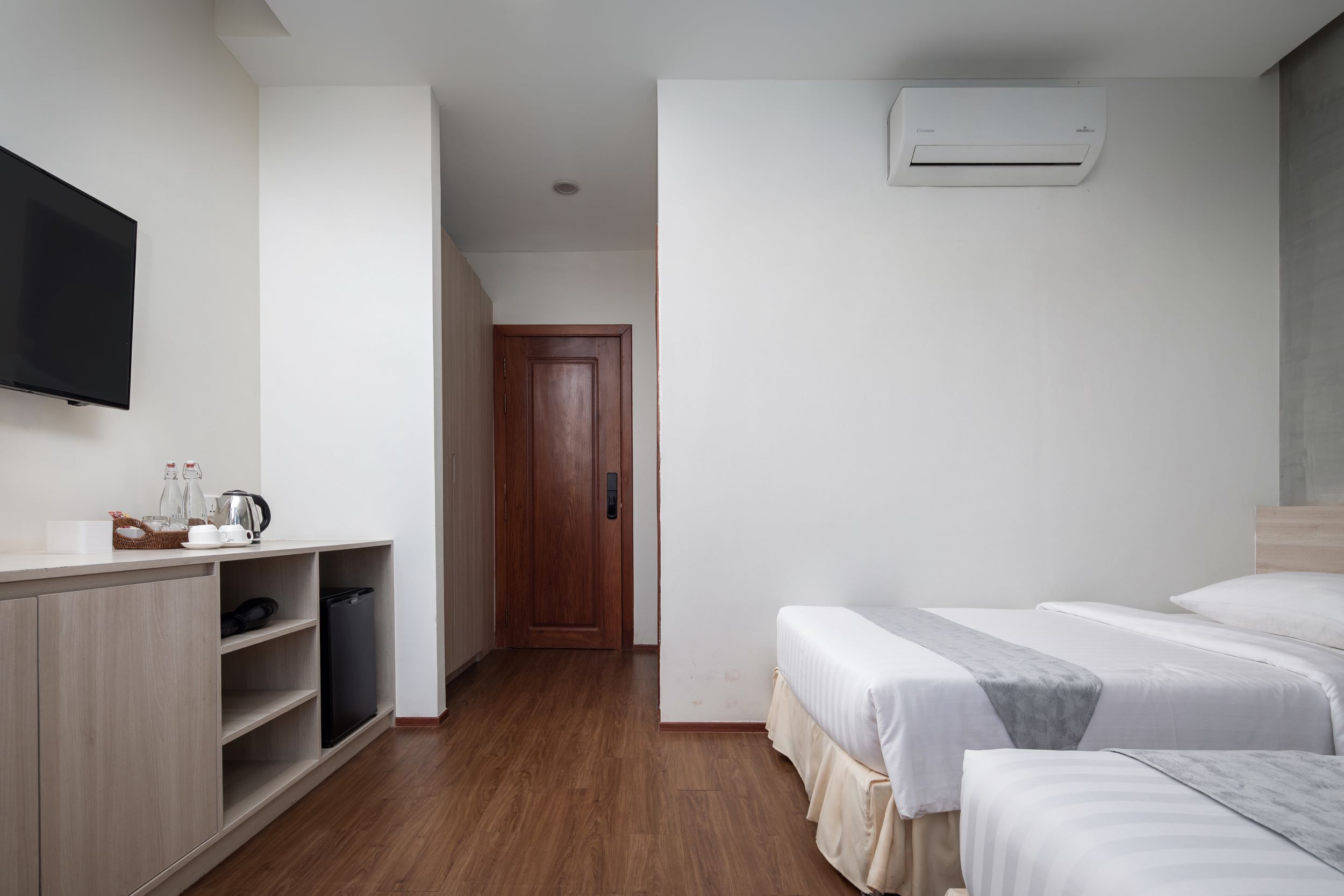 A modern hotel room with a wooden floor, a flat-screen TV, a wardrobe, a bed with white bedding, and an air conditioning unit.