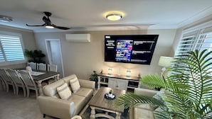 Een woonkamer met een bank, salontafel en een aan de muur bevestigde televisie. Er is een eethoek met een tafel en stoelen.