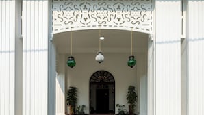 Entrada de un edificio con fachada blanca, un arco decorativo, plantas en macetas y un parterre circular con nenúfares.