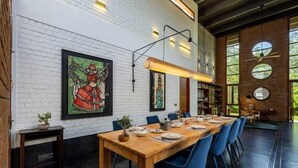 A dining area with a long wooden table set for six, blue chairs, and artwork on the white brick walls.