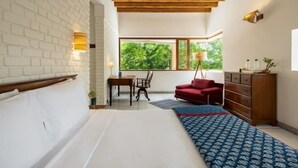 A bedroom with a bed, a red chair, a wooden dresser, a window with a view of greenery, and a white brick wall.