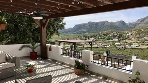 A covered outdoor seating area with wicker furniture, potted plants, and a view of a mountainous landscape.