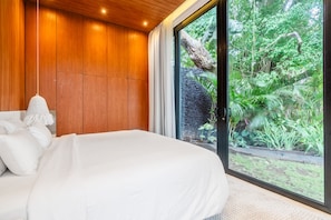 A bedroom with a large bed, wooden panel walls, and a view of a tropical garden through a sliding door. - Amazing 3BR Green Flow Villa in Ubud with Pool (Kec. Ubud)
