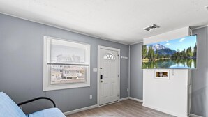 Una habitación con una cama, una ventana con persianas, una puerta y un televisor montado que muestra una escena de un lago de montaña.