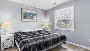 Un dormitorio con cama, dos mesitas de noche con lámparas, una ventana con persianas y un cuadro en la pared.