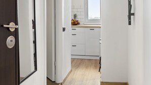 Un couloir doté d’un plancher en bois mène à une cuisine avec des meubles blancs et une fenêtre.