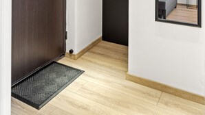 Un couloir avec une porte en bois, une porte noire et un miroir qui reflète un lit et une plante.