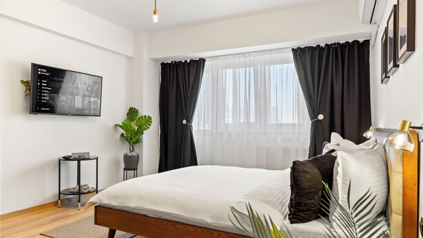 Une chambre moderne avec un lit en bois, un téléviseur fixé au mur, une plante en pot et une grande fenêtre ornée de rideaux légers.