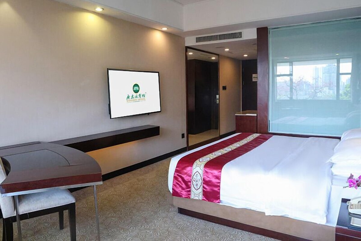 A hotel room with a bed, a desk, a chair, a TV mounted on the wall, and a window.