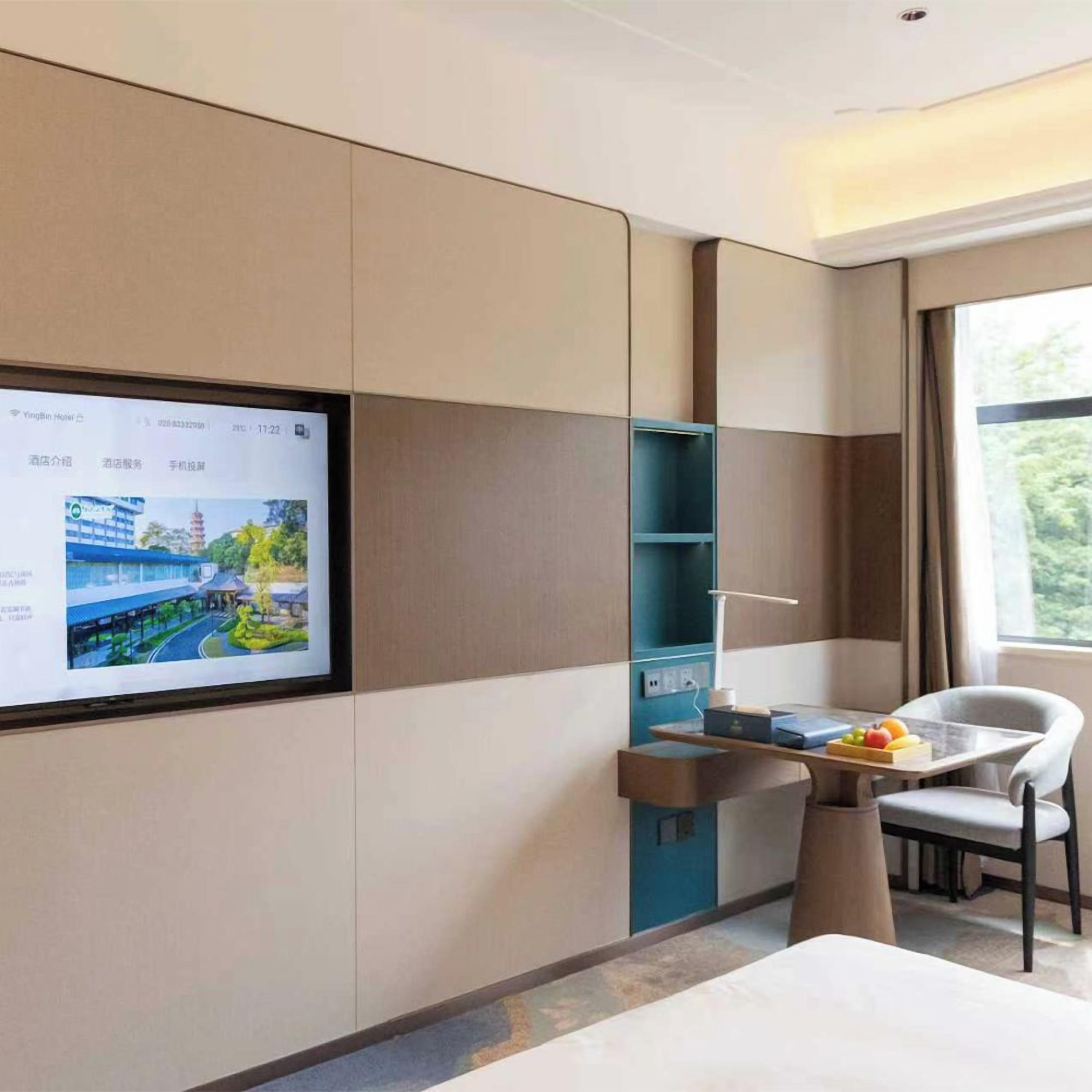 A hotel room with a large digital screen displaying a hotel map, a desk with a chair, and a window with a view of greenery.
