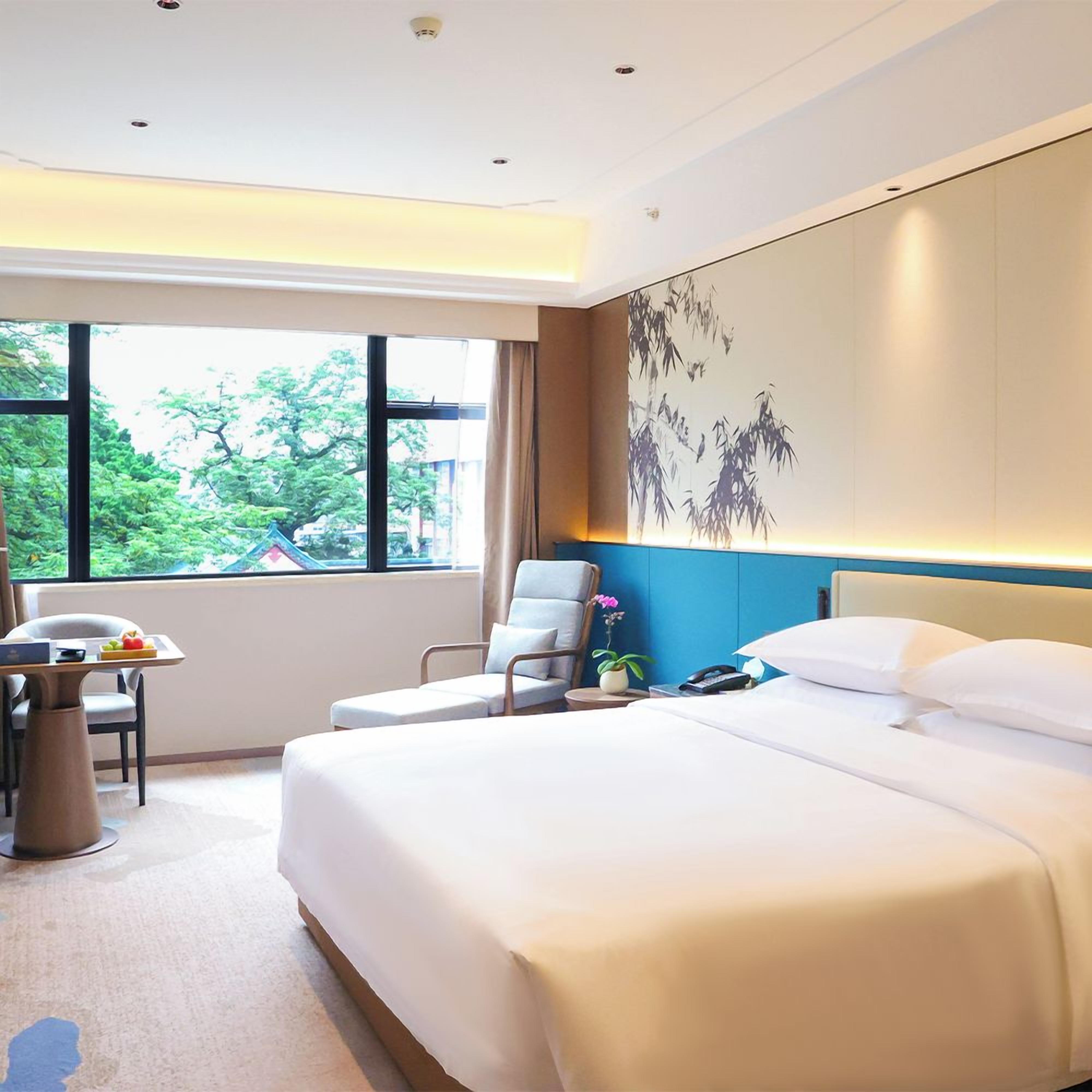 A hotel room with a large bed, a desk, a chair, and a view of greenery outside.