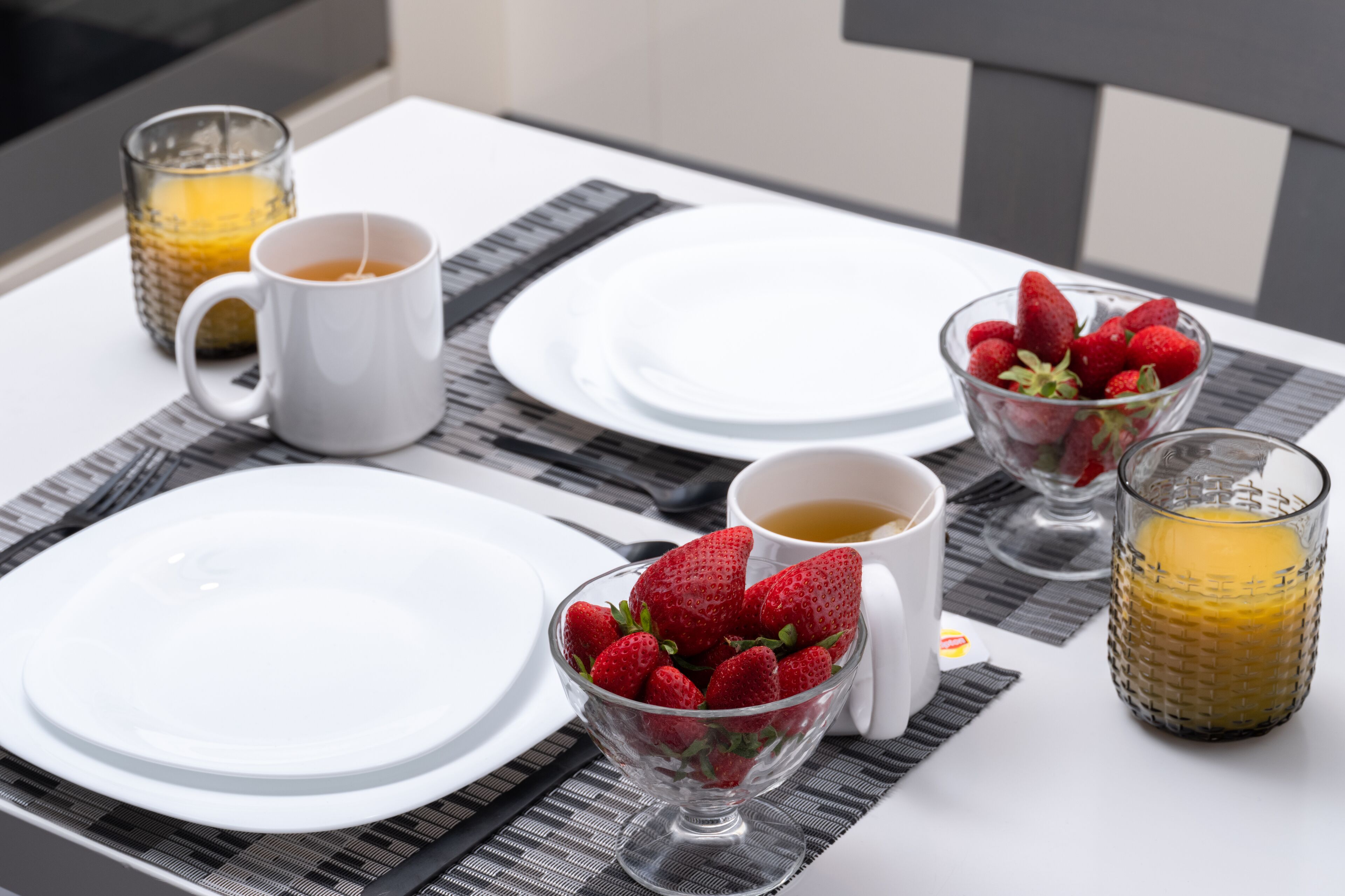 Une table mise pour le petit-déjeuner avec des assiettes blanches, un bol de fraises et deux verres de jus d’orange, le tout posé sur une table recouverte de sets de table noirs et blancs.