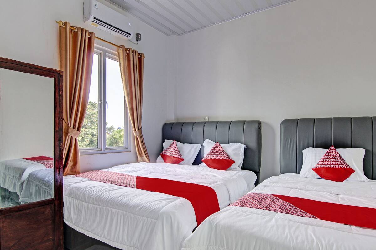 A hotel room with two beds, each with white bedding and red accents, a large mirror, and a window with curtains.
