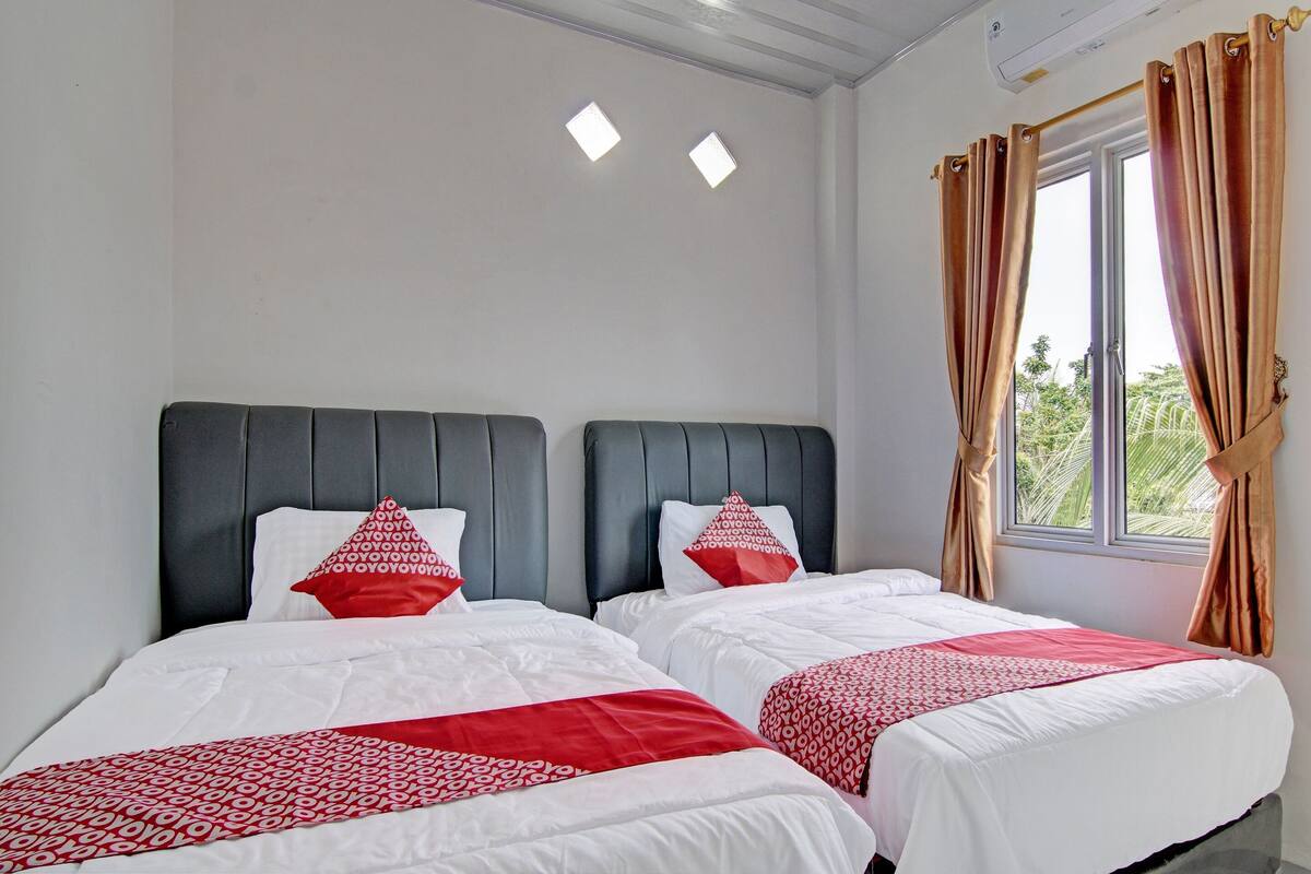 A hotel room with two beds, each with white bedding and red accents, a dark headboard, and a window with curtains.