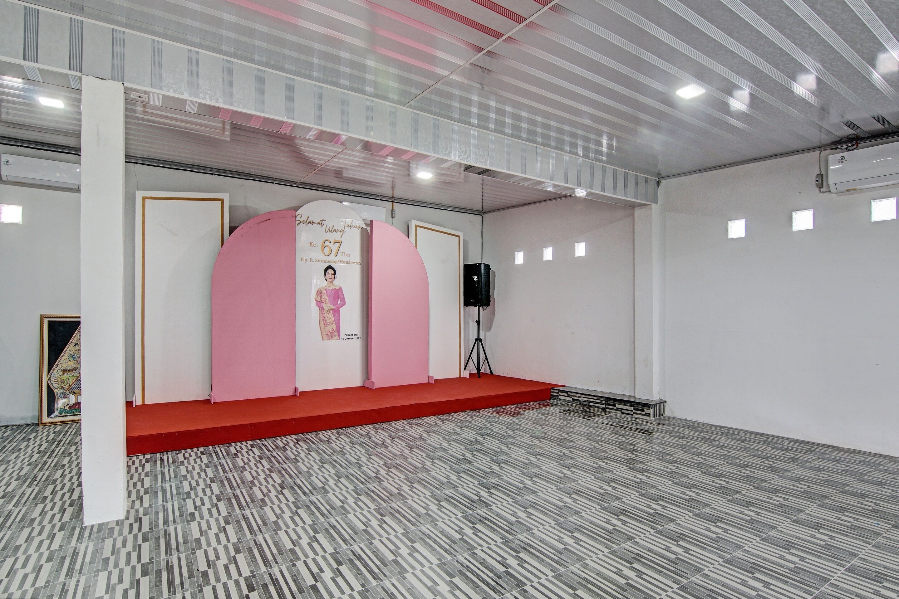 Um espaço amplo com palco revestido de carpete vermelho, paredes brancas e piso estampado.