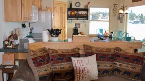 A cozy kitchen with wooden cabinets, a dining area with chairs, and a window with a view of trees. - Marr's Mountain Cabins - Cabin 1 (Red Feather Lakes)