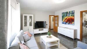 Un salon moderne avec un canapé gris, une table basse blanche, un téléviseur à écran plat et un coin cuisine doté de placards et d’un tableau.
