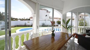 Un espace repas avec une table et des chaises en bois, de grandes fenêtres et une vue sur une piscine et un jardin.