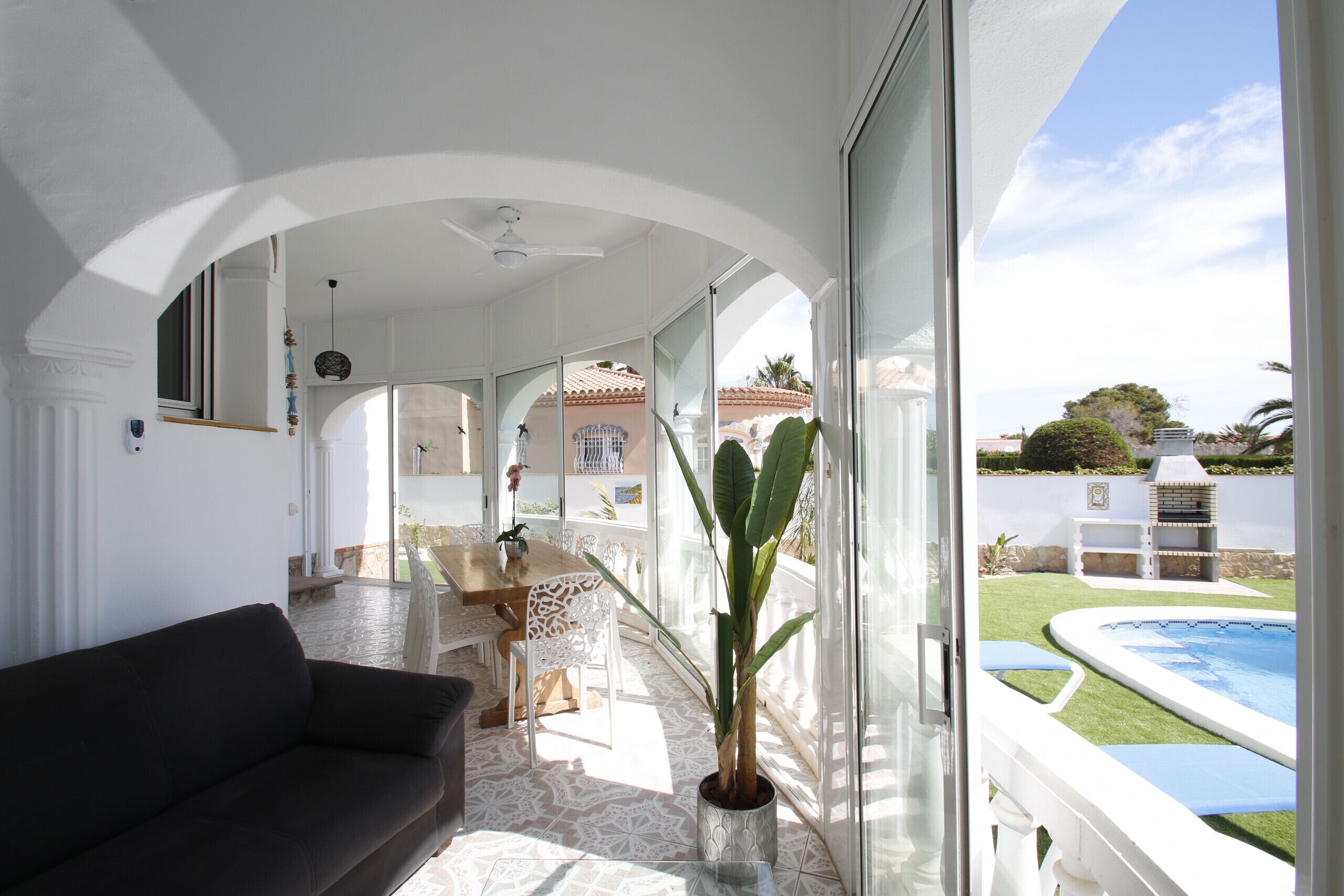 A spacious living area with a dark-colored sofa, a dining table with chairs, a potted plant, and a view of a swimming pool and garden.