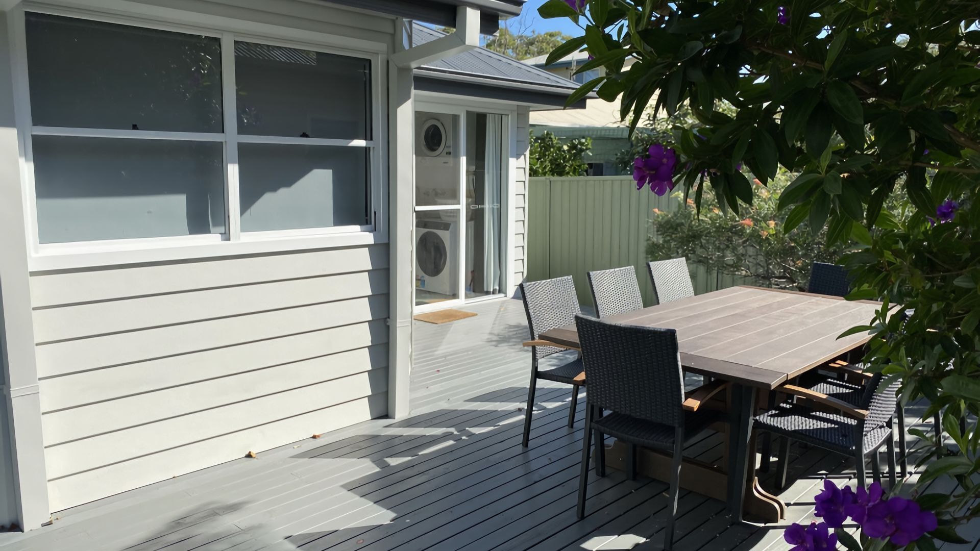 Een overdekt terras met een houten terras, een tafel met stoelen en een kleine tuin met paarse bloemen.