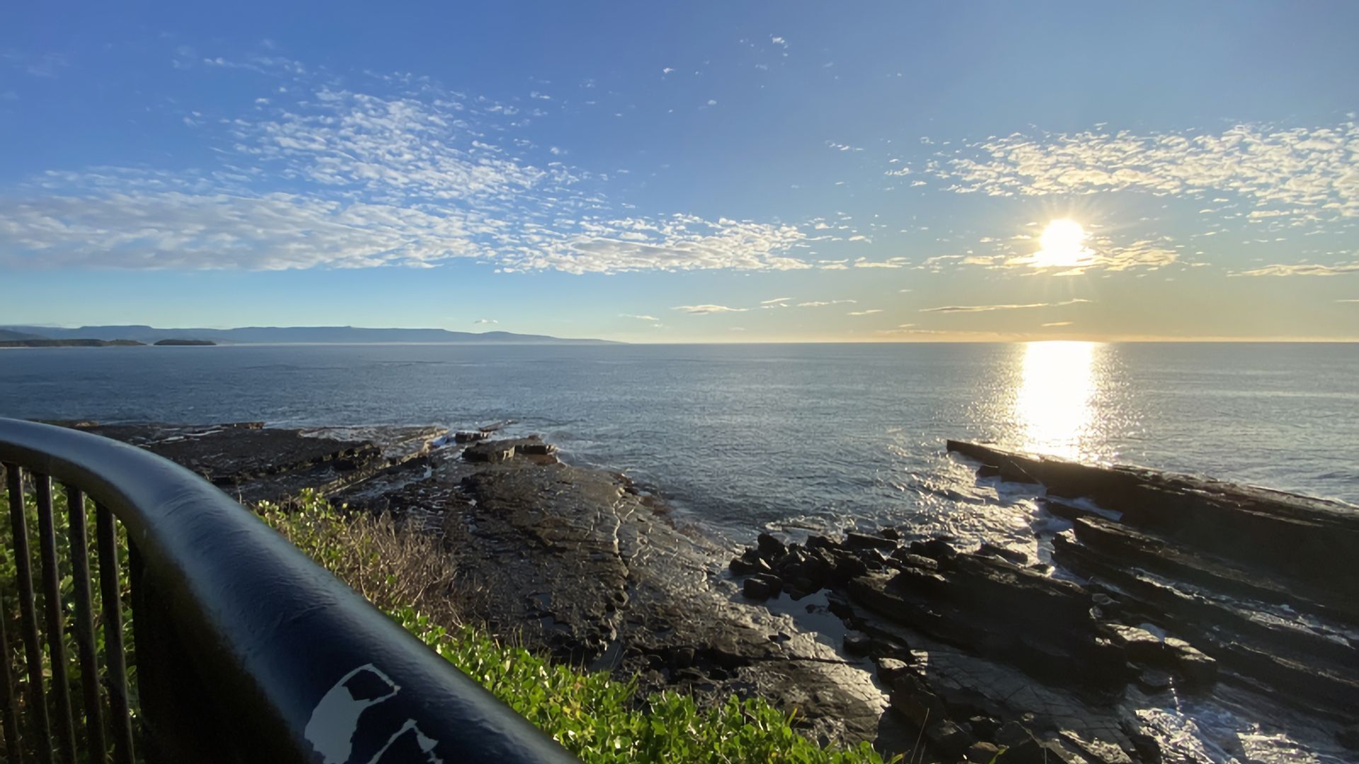 Vista costera con el sol poniéndose sobre el agua, costa rocosa y barandilla de metal.